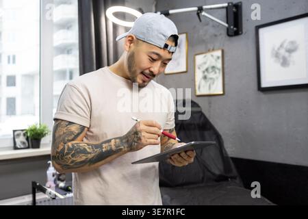 Ein lächelnder asiatischer Tätowierer, der auf Tablet neue Kunstideen ausarbeitet, während er in seinem Tätowierstudio steht Stockfoto