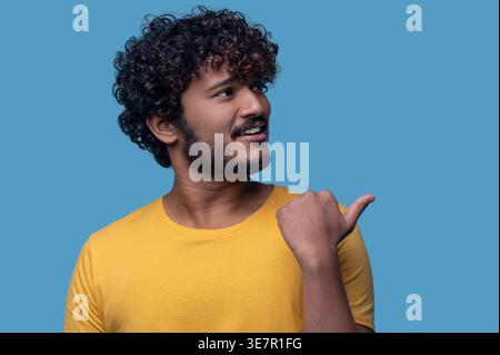 Lächelnder indischer Mann mit lockigem Haar, der wegschaut, mit Daumen, der seitlich vor blauem Hintergrund zeigt Stockfoto