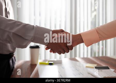 Asiatische Designerin und Kollegin schüttelt sich die Hand, während sie im Büro an den Tischunterlagen steht Stockfoto