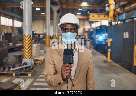 Schwarzer männlicher Ingenieur mit Schutzhelm und Gesichtsmaske, der das Mikrofon im Lager hält Stockfoto