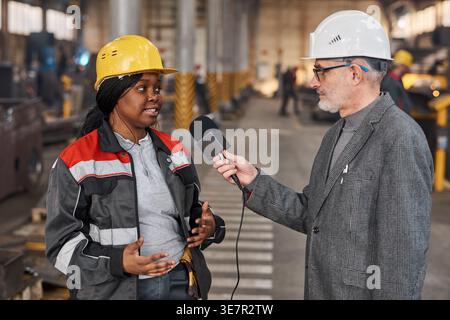 Schwarze Arbeitnehmerin mit Schutzhelm, die einen kaukasischen Mann im Lager befragt Stockfoto