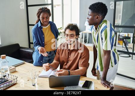 Mann aus dem Nahen Osten, der am Holztisch sitzt und einen neuen Projektplan bespricht, mit schwarzen männlichen und weiblichen Kollegen, die neben ihm im Büro stehen Stockfoto