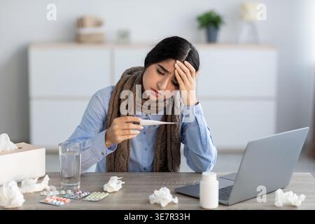 Kranke indische Freiberuflerin, die einen Schal trägt, der mit einer Hand den Kopf hält und die Temperatur mit einem Thermometer am Tisch mit Laptop und Medi... Stockfoto