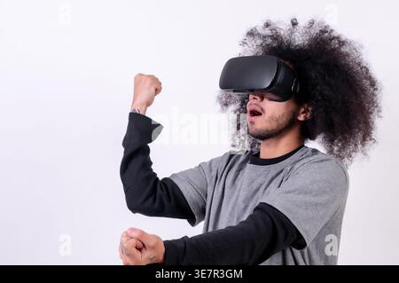 Hispanischer Mann mit lockigem Haar, der im Studio eine VR-Brille mit geballten Fäusten vor weißem Hintergrund trägt Stockfoto