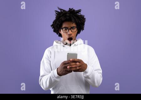 Schockierte den Schwarzen Mann mit Afro, der weißen Hoodie und Brille mit dem Smartphone trug, während er im Studio gegen die violette Wand stand Stockfoto