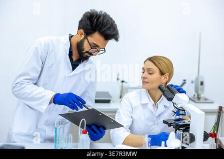 Indischer Wissenschaftler mit Labormantel und Latexhandschuhen macht Notizen, nachdem er einen Probentest in der Nähe einer kaukasischen Kollegin gemacht hat, die ein Mikroskop in La... Stockfoto