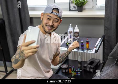 Lächelnder asiatischer Tätowierer, der Selfie auf dem Smartphone macht, während er in seinem Tätowierstudio sitzt Stockfoto