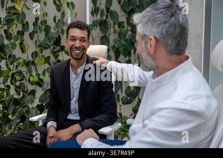 Ein lächelnder Schwarzer Mann, der mit einem männlichen kaukasischen Psychotherapeuten spricht, der in der Klinik auf einem Stuhl sitzt Stockfoto