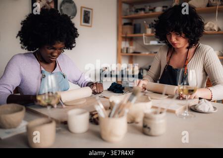 Schwarze Künstlerin mit Afroschürze, die mit einem Rollstift auf Lehm sitzt, während sie neben der Co-Künstlerin im Studio sitzt Stockfoto