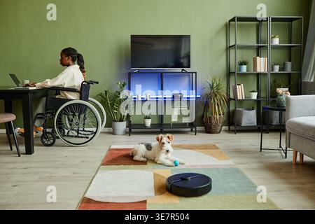 Behinderte Schwarze Frau im Rollstuhl sitzt am Tisch, arbeitet an einem Laptop in der Nähe ihres Haustierhundes, der zu Hause auf Teppich sitzt Stockfoto