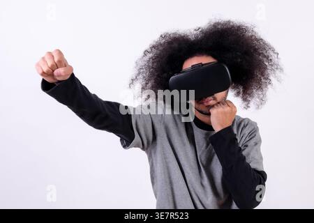 Hispanischer Mann mit lockigem Haar, der im Studio eine VR-Brille mit geballten Fäusten vor weißem Hintergrund trägt Stockfoto