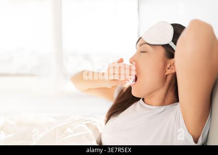 Asiatische Frau gähnt und dehnt sich im Bett mit Augenmaske auf der Stirn Stockfoto
