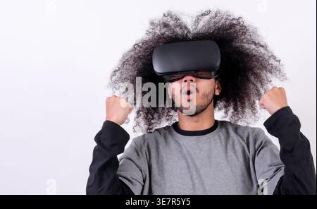 Hispanischer Mann mit lockigem Haar, der im Studio eine VR-Brille mit geballten Fäusten vor weißem Hintergrund trägt Stockfoto