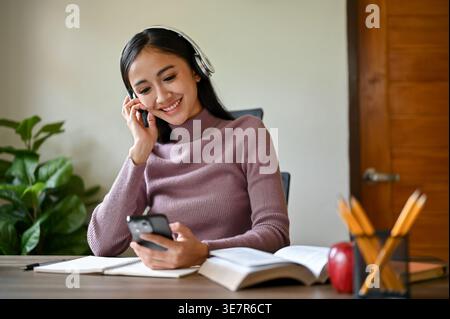 Lächelnde asiatische Frau im Kopfhörer, die Musik mit dem Smartphone hört, während sie zu Hause mit Büchern am Tisch sitzt Stockfoto