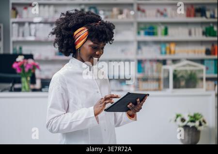 Schwarze Apothekerin mit Afro-Haltung und Verwendung der Tablette, die in der Apotheke gegen den Zähler steht Stockfoto