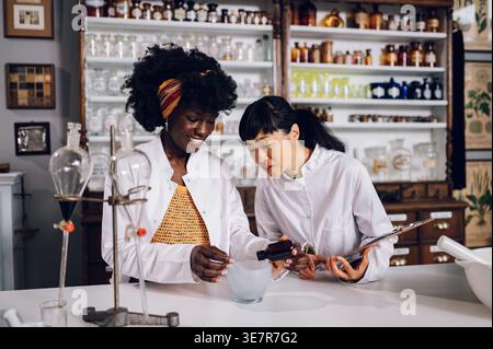 Schwarze Chemikerin gießt Zutat in Mörtel, während japanische Kollegin die stehende Holding Chart in Vintage-Apotheke beobachtet Stockfoto