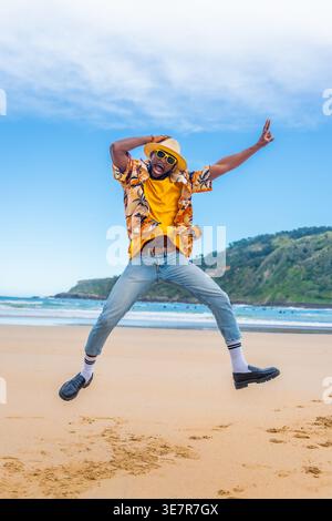 Schwarzer Mann mit hawaiianischem Hemd und Strohhut, der im Sommer mit erhobenen Armen am Strand springt Stockfoto