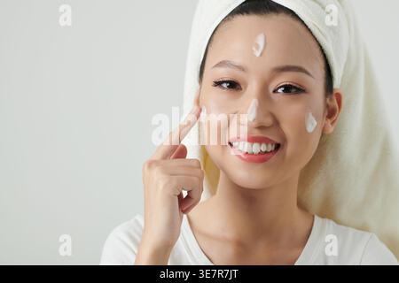 Glückliche asiatische Frau mit Handtuch um den Kopf gewickelt, die Gesichtscreme aufträgt, die vor weißem Hintergrund im Studio steht Stockfoto