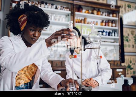 Fokussierte schwarze Chemikerin, die das Ventil des Trenntrichters öffnet und es in den Becher fließen lässt, der mit einer japanischen Kollegin in in Vintage... Stockfoto