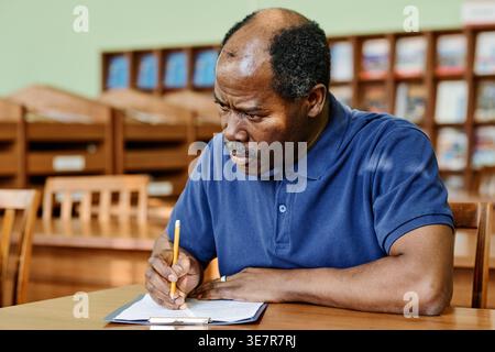 Schwarzer, männlicher Immigrant mittleren Alters, der am Schreibtisch sitzt und einen englischen Grammatiktest in der Bibliothek absolviert Stockfoto