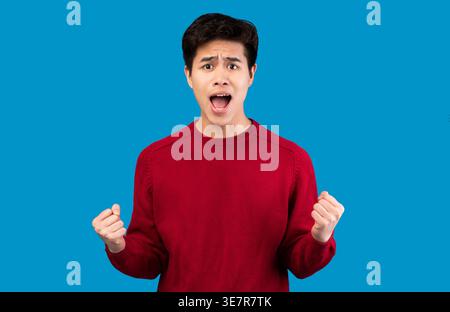Asiatischer Mann mit rotem Pullover, der schreiend mit Fäusten vor der blauen Wand im Studio steht Stockfoto