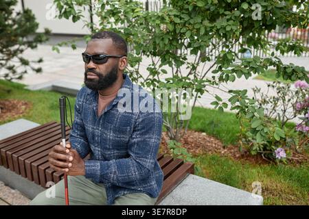 Behindertengerechter Schwarzer Mann mit Sonnenbrille und Gehstock, der tagsüber auf einer Bank im Stadtpark sitzt Stockfoto