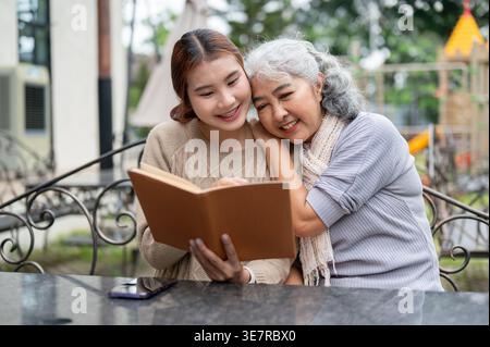 Thai Senior Frau, die Spaß beim Lesen von Büchern mit ihrer Enkelin hat, während sie tagsüber am Tisch im Garten sitzt Stockfoto