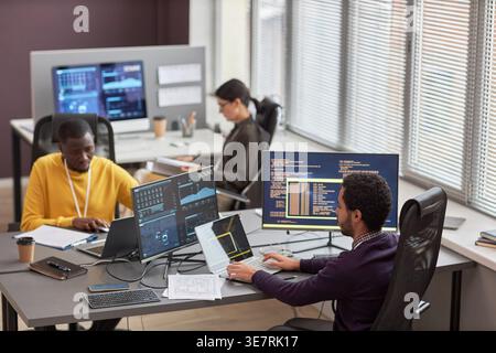 Schwarze und gemischte männliche Programmierer arbeiten an einem Laptop in der Nähe einer kaukasischen Kollegin, während sie am Tisch sitzen und im Büro mit Computern sitzen Stockfoto