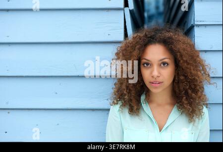 Ernsthafte gemischte Rassenfrau mit lockigen Haaren, die weiße lange Ärmel trägt und draußen an der blauen Wand steht Stockfoto