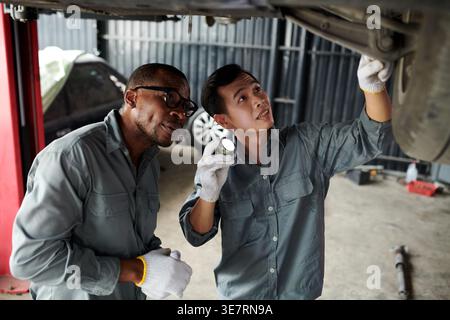 Schwarze und asiatische männliche Mechaniker mit Handschuhen, die unter dem Fahrgestell des Fahrzeugs arbeiten, während sie in der Autowerkstatt stehen Stockfoto