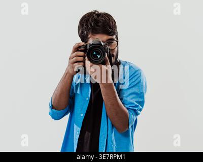 Indischer Mann in blauen langen Ärmeln mit Brille, die Bilder mit DSLR-Kamera macht, während er im Studio vor weißem Hintergrund steht Stockfoto