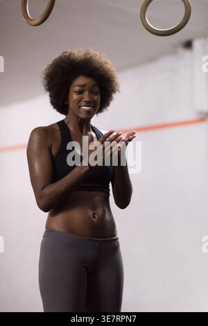 Fröhliche schwarze Frau mit Afro-Kleidung, die ihre Hände überprüft, nachdem sie im Fitnessstudio Ringtauchtraining gemacht hat Stockfoto