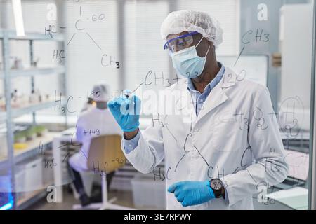 Schwarzer Wissenschaftler mit Labormantel und Maske, der chemische Formeln auf Glasplatte schreibt, während er in der Nähe eines männlichen Kollegen im Labor steht Stockfoto