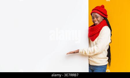 Lächelnde schwarze Frau mit roter Mütze und Schal, die leeres Whiteboard vor gelbem Hintergrund im Studio zeigt Stockfoto