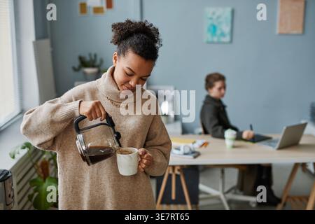 Geschäftsfrau mit gemischter Rasse, die Kaffee aus der Teekanne in eine Tasse gießt, während sie in der Pause im Büro neben einer kaukasischen Kollegin steht Stockfoto