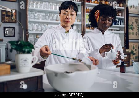 Japanische Apothekerin, die Tee packt, steht mit schwarzer Kollegin, die Medizin in Mörtel in Vintage-Apotheke mischt Stockfoto