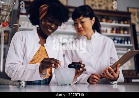 Schwarze Chemikerin gießt Zutat in Mörtel, während japanische Kollegin die stehende Holding Chart in Vintage-Apotheke beobachtet Stockfoto