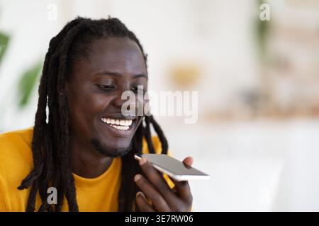 Ein lächelnder Schwarzer Mann mit Dreadlocks, der Sprachnachrichten aufnimmt, sitzt auf dem Sofa im Wohnzimmer zu Hause Stockfoto