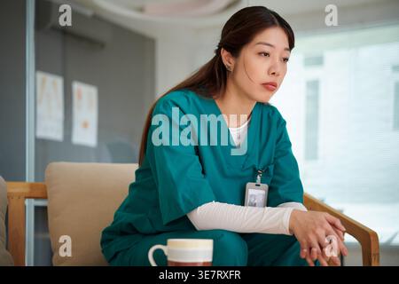 Asiatische Krankenschwester trägt einen Scheueranzug und blickt weg, während sie mit gefalteten Händen auf dem Sofa in der Krankenhauslounge sitzt Stockfoto