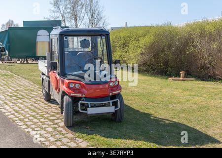Rotes Nutzfahrzeug auf Grasfeld geparkt. Klarer blauer Himmel über grünen Büschen. Das Fahrzeug verfügt über eine Schutzkabine mit Fenstern. Keine Leute sichtbar, Szene Appeal Stockfoto