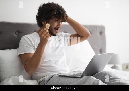 Gestresster bärtiger indischer Mann, der lockiges Haar berührt und goldenes Bitcoin hält, der auf dem Bett zu Hause sitzt Stockfoto