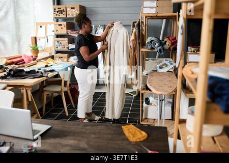 Serious Black Modedesignerin steht und misst unvollendete Mäntel an Schaufensterpuppe mit Bandmaß in der Schneiderei Stockfoto