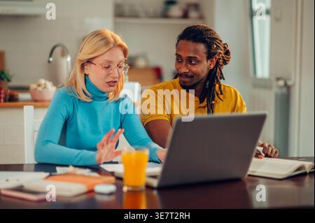 Ein lächelnder Mixed Race Mann mit Schlössern, der die kaukasische Freundin ansieht, die ihre Rechnungen und Steuern berechnet und zu Hause mit Laptop am Tisch sitzt Stockfoto