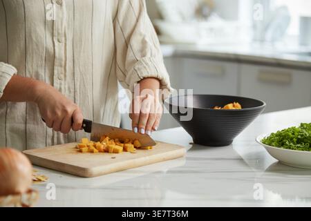 Hände einer Frau mit gemischter Rasse, die Kürbis mit einem Messer auf Schneidebrett schneidet und tagsüber auf der Küchenarbeitsplatte steht Stockfoto