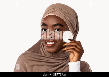 Lächelnde schwarze muslimische Frau, die Hijab trägt und Toner mit Wattepad auf das Gesicht aufträgt, während sie mit vor weißem Hintergrund im Studio steht Stockfoto