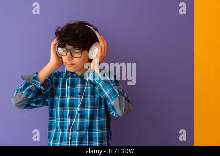 Ernster männlicher Teenager aus dem Nahen Osten in karierten langen Ärmeln und Brillen, der Musik über Kopfhörer hört und wegblickt, während er im Stehen steht... Stockfoto