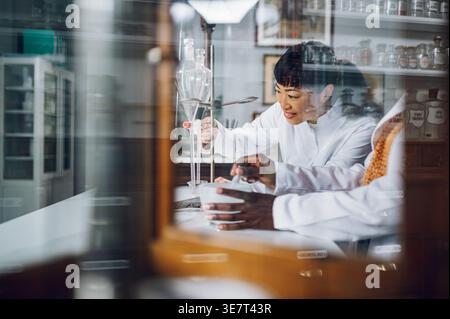 Japanische Chemikerin im Laborkittel, die sich auf das Gegenöffnungsventil des Trenntrichters stützt und es in das Becher fließen lässt, das neben Blac steht... Stockfoto