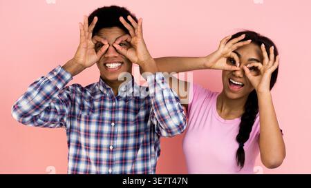 Lächelndes schwarzes Paar macht Handbrillen, während es im Studio vor rosa Hintergrund steht Stockfoto