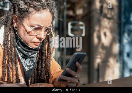 Hispanische Frau mit Dreadlocks, die eine Brille mit Smartphone trägt und tagsüber am Tisch mit Kaffeetasse im Café im Freien sitzt Stockfoto