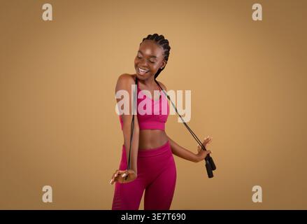 Fröhliche schwarze Frau in Fitnesskleidung, die ein Sprungseil um ihre Schultern hält, während sie im Studio vor braunem Hintergrund steht Stockfoto